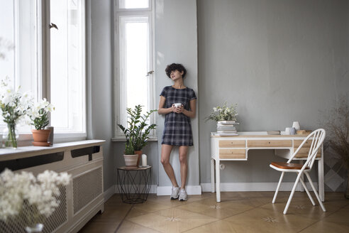 Junge Frau mit Kaffeetasse schaut durch das Fenster zu Hause - RBF05512