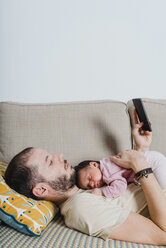 Vater auf der Couch liegend mit neugeborenem kleinen Mädchen und Smartphone - GEMF01351