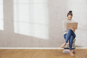 Junge Frau sitzt auf einem Stapel von Büchern und benutzt einen Laptop zu Hause - FMKF03400