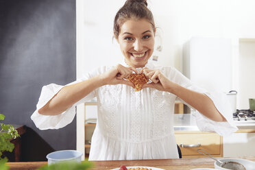 Porträt einer jungen Frau mit herzförmiger Waffel in der Küche - FMKF03414