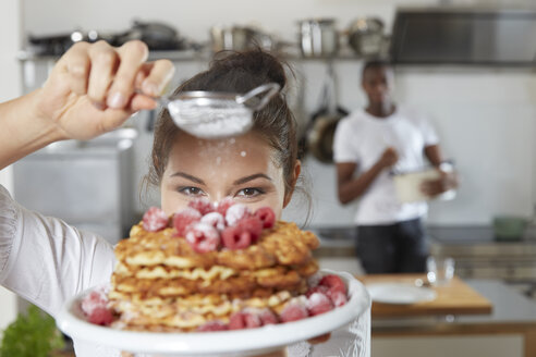 Portrait of young woman sprinkling icing sugar on waffles in the kitchen - FMKF03416
