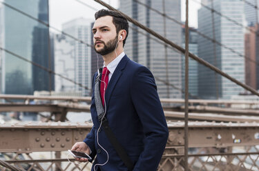 USA, New York City, businessman with cell phone and earbuds on Brooklyn Bridge - UUF09632