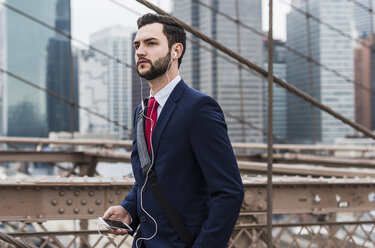 USA, New York City, Geschäftsmann mit Mobiltelefon und Kopfhörern auf der Brooklyn Bridge - UUF09632