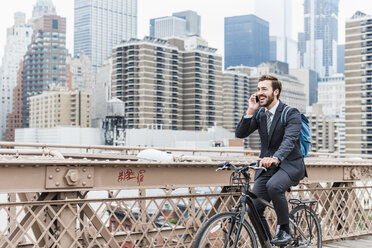 USA, New York City, laughing businessman on bicycle on Brooklyn Bridge using cell phone - UUF09640