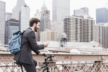 USA, New York City, businessman on bicycle on Brooklyn Bridge using cell phone - UUF09642