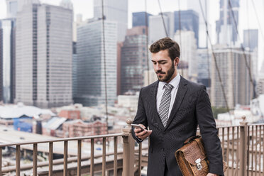 USA, New York City, Geschäftsmann auf der Brooklyn Bridge, der ein Mobiltelefon benutzt - UUF09652