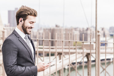 USA, New York City, lächelnder Geschäftsmann mit Handy und Kopfhörern auf der Brooklyn Bridge - UUF09653