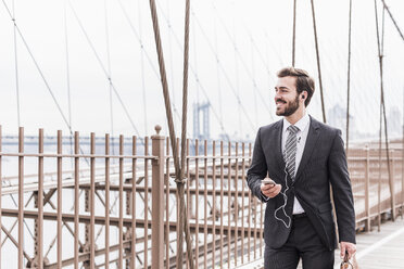 USA, New York City, lächelnder Geschäftsmann mit Handy und Kopfhörern auf der Brooklyn Bridge - UUF09654