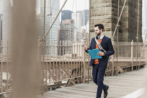 USA, New York City, Mann geht auf der Brooklyn Bridge und benutzt ein Mobiltelefon, lizenzfreies Stockfoto
