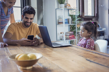Familie sitzt in der Küche, Eltern benutzen ein Smartphone, Tochter schaut auf den Laptop - WESTF22445