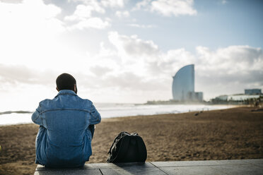 Spain, Barcelona, back view of young traveler sitting at the beach - JRFF01139