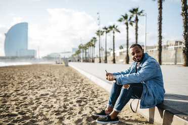 Spain, Barcelona, smiling young traveler sitting at the beach - JRFF01140