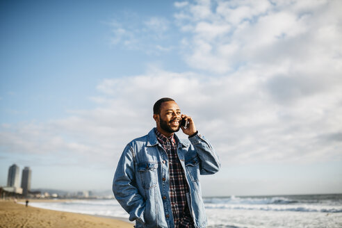 Spanien, Barcelona, lächelnder junger Mann mit Mobiltelefon am Strand - JRFF01145