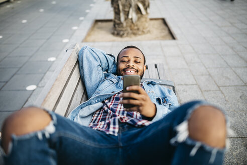 Porträt eines lächelnden jungen Mannes, der auf einer Bank liegt und auf sein Mobiltelefon schaut - JRFF01147