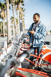 Spain, Barcelona, young man with smartphone renting a city bike - JRFF01154