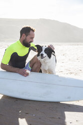 Mann mit Hund, der sein Surfbrett am Strand mit Paraffin einölt - ABZF01708