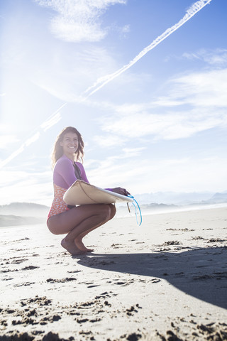 Lächelnde Frau mit Surfbrett am Strand hockend, lizenzfreies Stockfoto