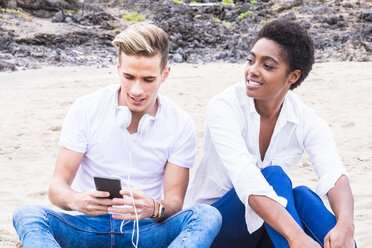 Junger Mann sitzt neben seiner Freundin am Strand mit Handy und Kopfhörern - SIPF01253
