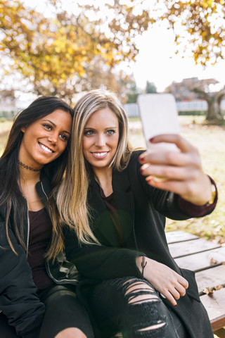 Zwei lächelnde junge Frauen auf einer Parkbank machen ein Selfie, lizenzfreies Stockfoto