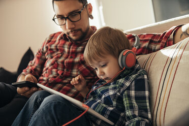 Father and son on couch using tablet and wearing headphones - ZEDF00502