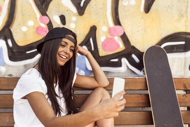Lächelnde junge Frau mit Skateboard schaut auf ihr Handy - MGOF02765
