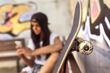 Skateboard und junge Frau mit Mobiltelefon im Hintergrund - MGOF02766
