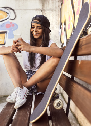 Lächelnde junge Frau mit Skateboard schaut auf ihr Handy, lizenzfreies Stockfoto