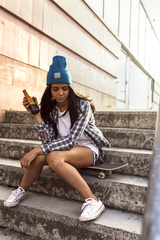 Junge Frau sitzt auf einem Skateboard und hält eine Bierflasche, lizenzfreies Stockfoto