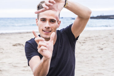 Porträt eines lächelnden jungen Mannes am Strand, der mit seinen Fingern einen Rahmen baut - SIPF01270