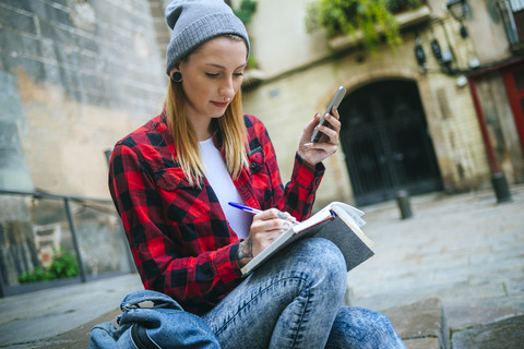 Spanien, Barcelona, junge Frau mit Handy sitzt auf einer Treppe und schreibt in ein Notizbuch, lizenzfreies Stockfoto