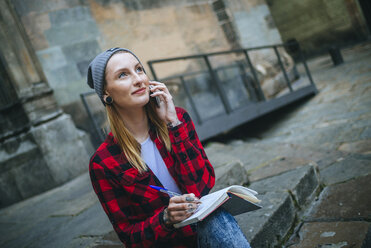 Spanien, Barcelona, Porträt einer lächelnden jungen Frau, die auf einer Treppe sitzend mit einem Notizbuch telefoniert - KIJF01056