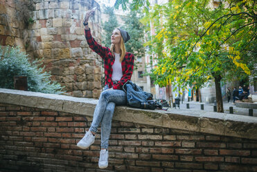 Spanien, Barcelona, junge Frau sitzt auf einer Mauer und macht ein Selfie mit Handy - KIJF01067