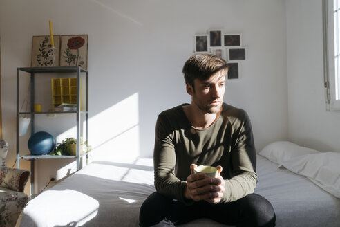 Unhappy young man sitting on bed with cup of coffee - KKAF00289