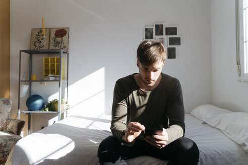 Young man sitting on bed looking at his smartphone - KKAF00290