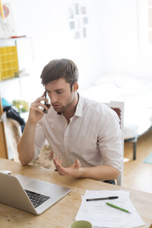 Junger Mann am Telefon, der von zu Hause aus mit Laptop arbeitet - KKAF00302