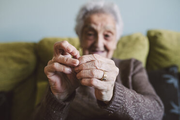 Hände einer älteren Frau, die auf einer Couch sitzt und den Faden durch das Knopfloch einer Nähnadel führt - RAEF01632