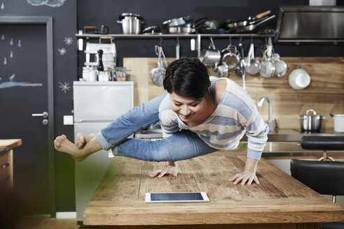 Frau macht einen Handstand auf dem Tisch in der Küche und schaut auf ein Tablet - FMKF03459