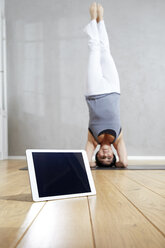 Woman practising yoga doing a headstand behind tablet - FMKF03471