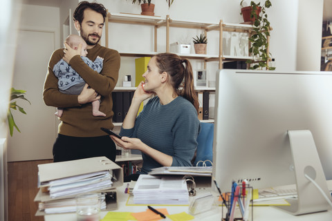 Lächelnde Mutter am Schreibtisch mit Blick auf den Vater, der das Baby im Büro zu Hause hält, lizenzfreies Stockfoto