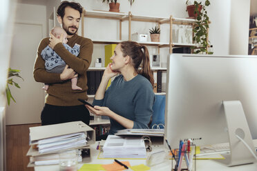 Lächelnde Mutter am Schreibtisch mit Blick auf den Vater, der das Baby im Büro zu Hause hält - MFF03425