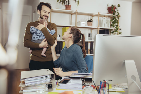 Lächelnde Mutter am Schreibtisch mit Blick auf den Vater, der das Baby im Büro zu Hause hält, lizenzfreies Stockfoto