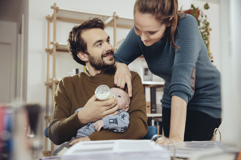 Mother looking at father feeding baby in home office - MFF03431