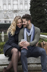 Spain, Madrid, happy couple with cell phone with the Royal Palace in background - ABZF01787