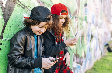 Zwei Kinder lehnen an einer Graffiti-Wand und benutzen Smartphones, lizenzfreies Stockfoto