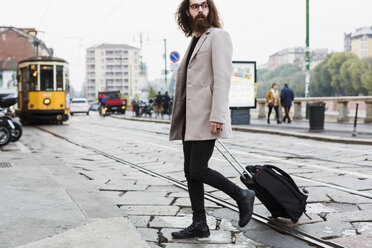 Stylish young man with suitcase crossing tramway - MAUF00929