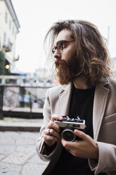 Stylish young man outdoors holding camera looking around - MAUF00939