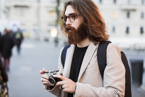 Stilvoller junger Mann im Freien, der eine Kamera hält und sich umschaut - MAUF00943