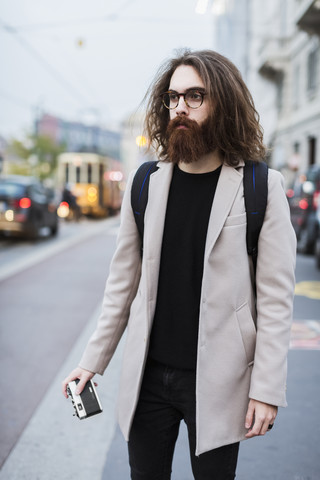 Stilvoller junger Mann in der Stadt, der eine Kamera hält und sich umschaut, lizenzfreies Stockfoto