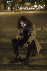 Stilvoller junger Mann im Freien in der Stadt bei Nacht, der auf sein Handy schaut - MAUF00972
