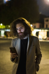 Stilvoller junger Mann im Freien in der Stadt bei Nacht, der auf sein Handy schaut - MAUF00973